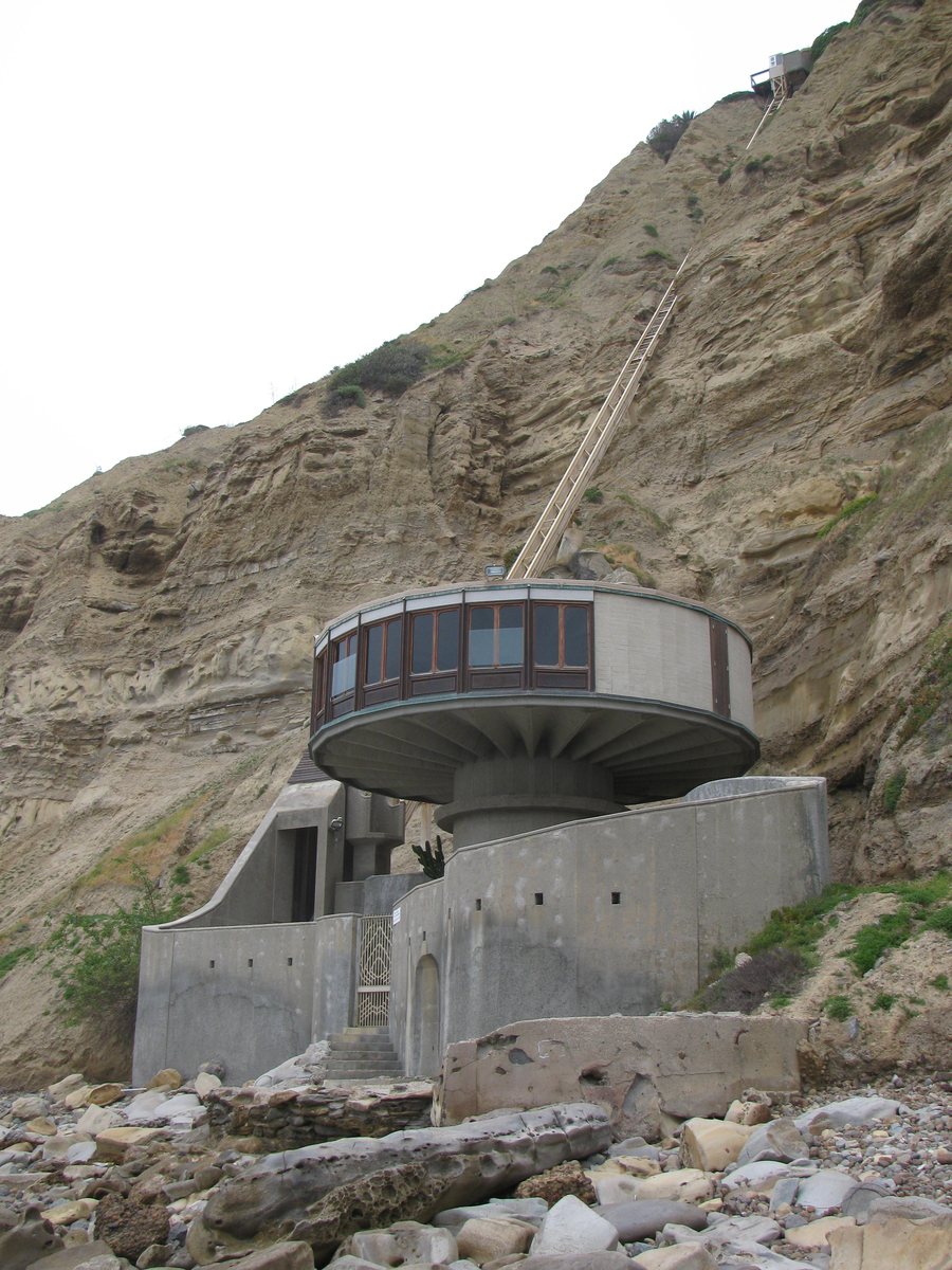 A round concrete house with a ring of windows sits on a pedestal at the foot of a steep sandstone cliff, behind a curved concrete wall above a rocky beach, with a narrow tram track climbing the cliff to a small landing at the top