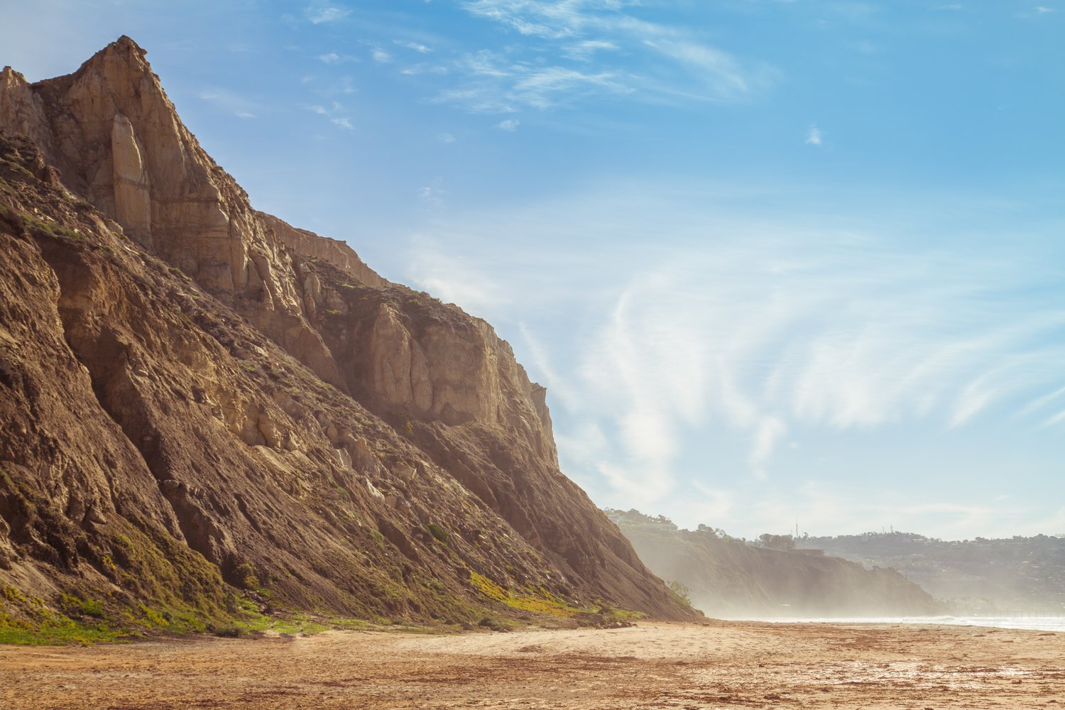 Tall eroded sandstone cliffs rise from a wide empty beach under a pale blue sky, with more bluffs fading into sea haze in the distance
