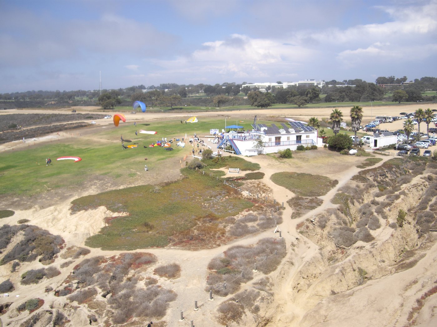 An aerial view of a grassy field on a cliff top with paragliders laid out on the grass, a small white clubhouse, palm trees and a parking lot, and eroded bluffs in the foreground