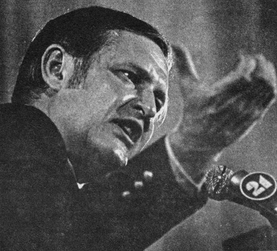 A black and white photograph of a man in a suit speaking into a television station microphone, gesturing with one hand