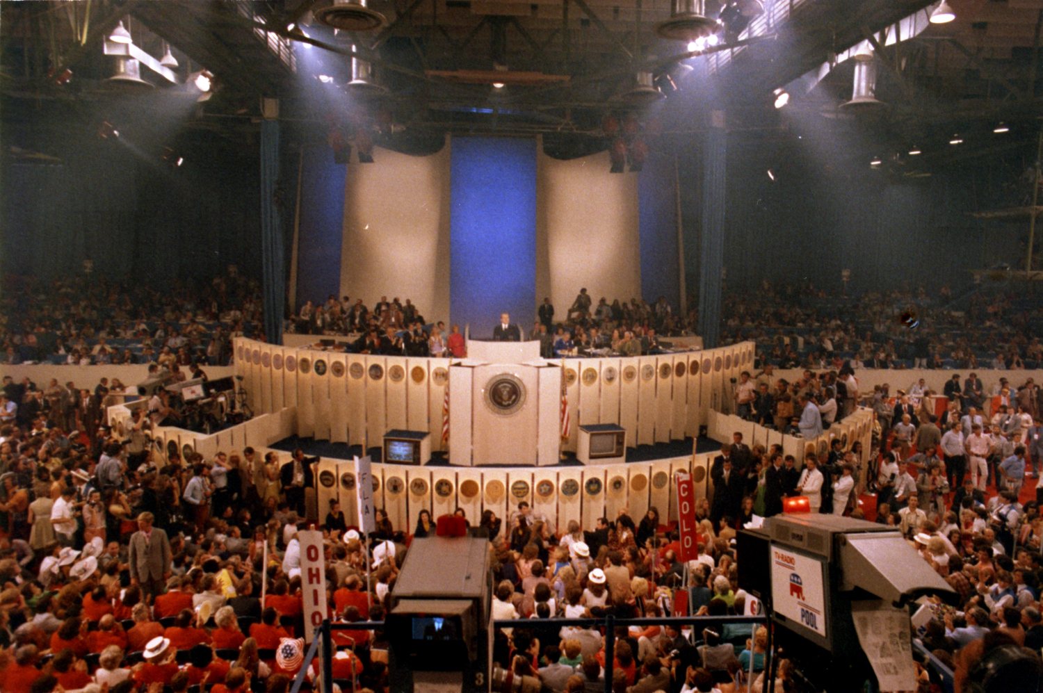 A packed convention hall seen from above, delegates with signs crowding the floor around a raised podium with a presidential seal and a blue backdrop