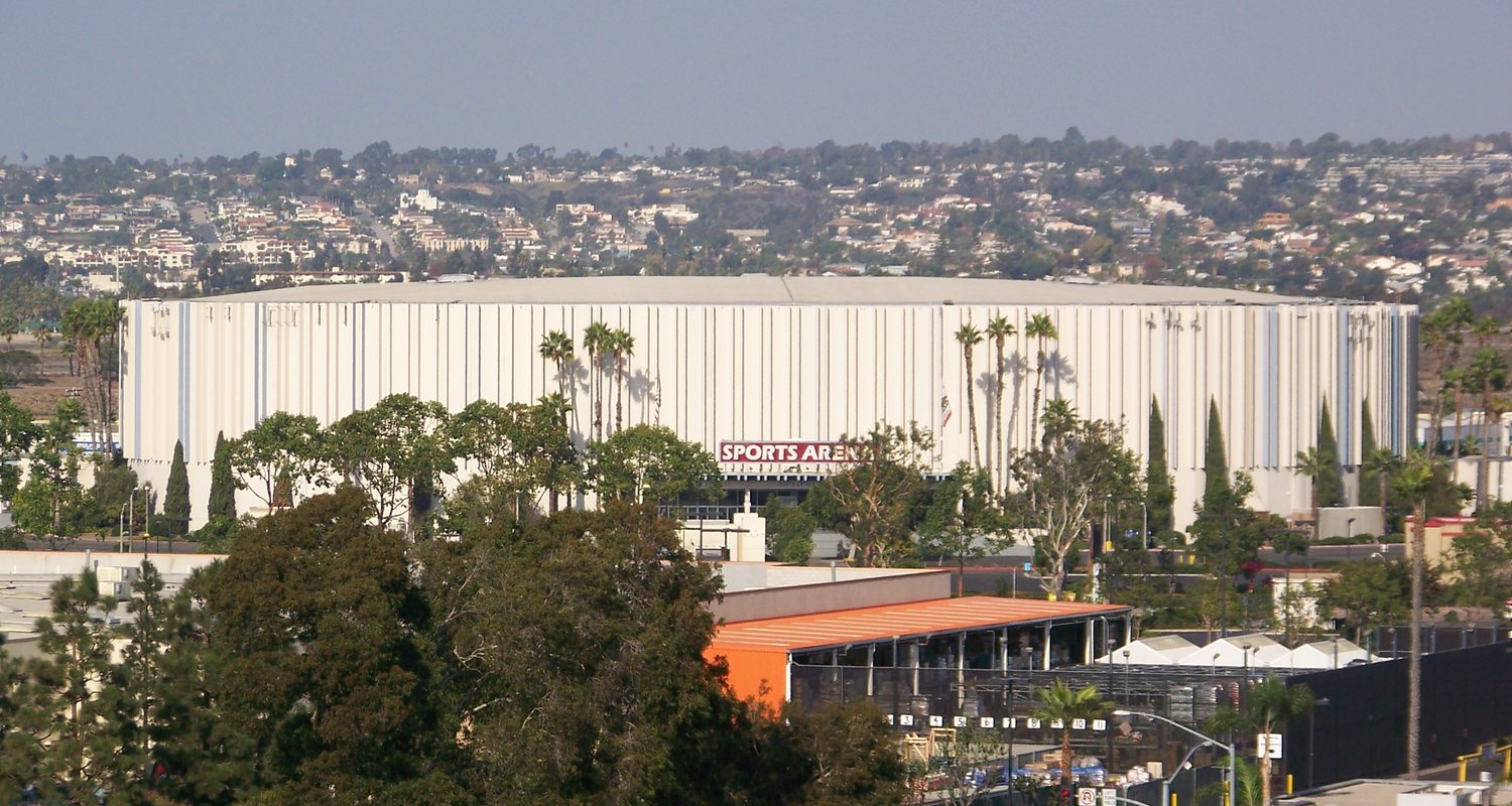 A long white arena building behind palm trees and a parking area, with hillside houses rising behind it