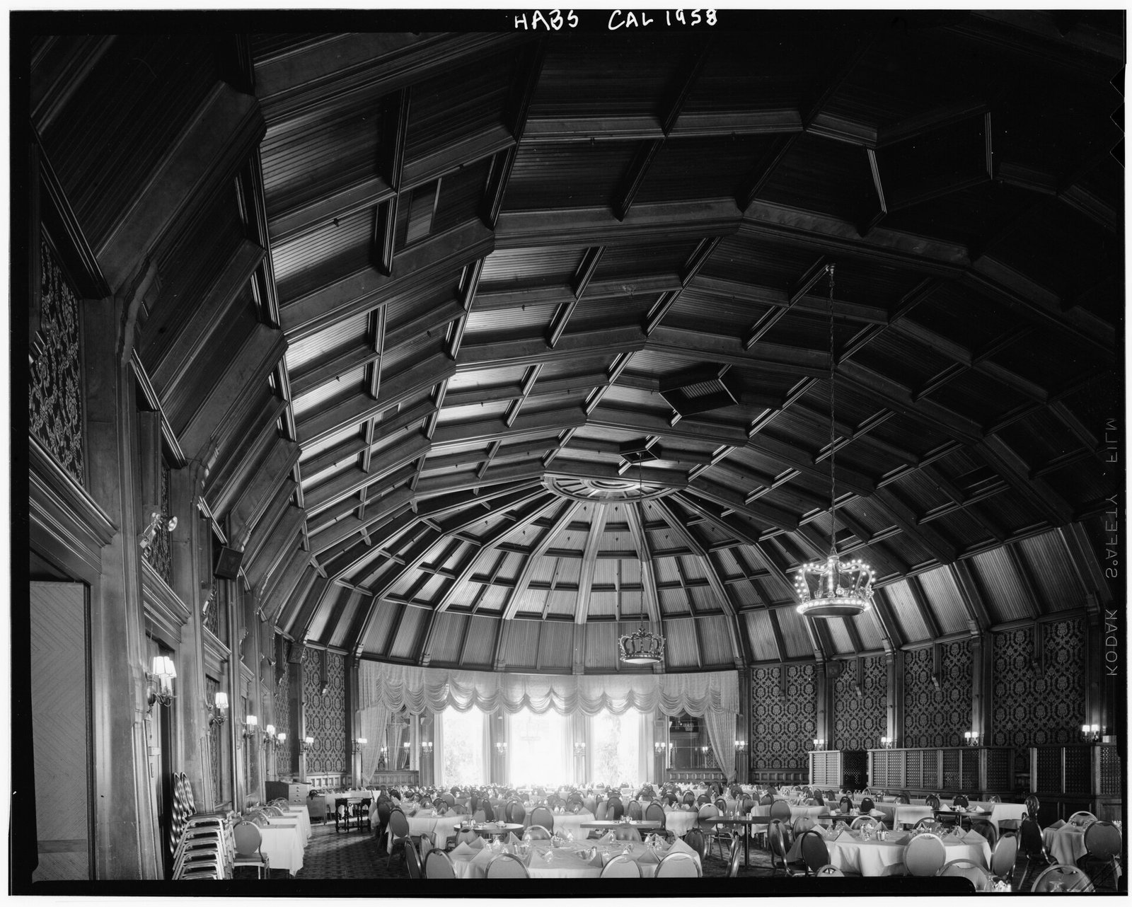 A long dining room under a high arched wooden ceiling of crossing ribs, tables set with white cloths and chandeliers hanging down