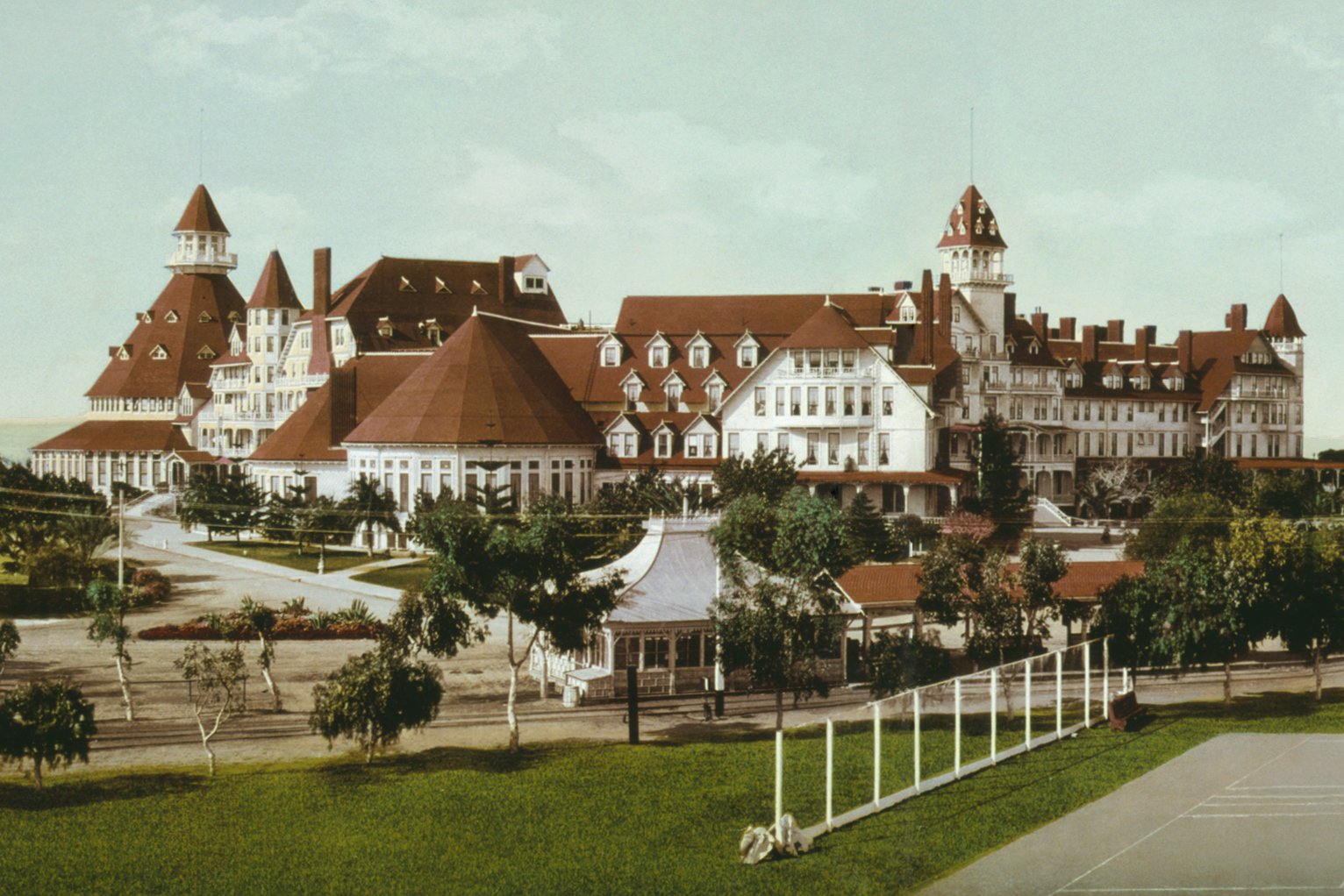 A color tinted view of the Hotel del Coronado, its red roofs, white walls and turrets behind lawns, palms and a tennis court