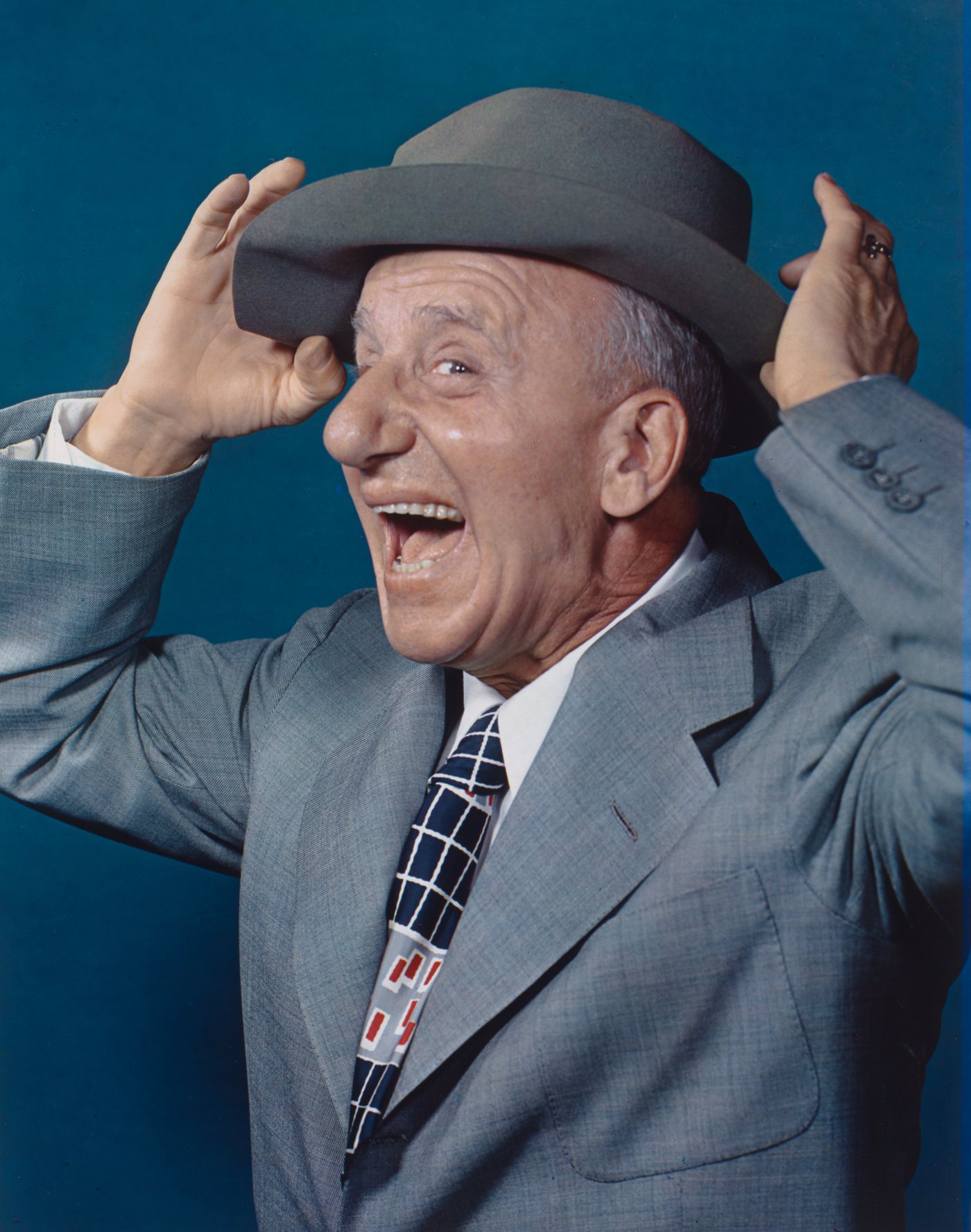 Color portrait of Jimmy Durante in a grey suit, tipping his hat back with both hands and laughing, photographed in 1948