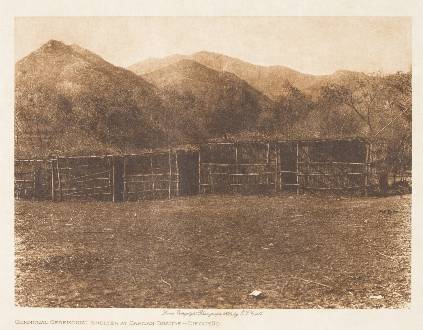 A long low shelter of brush and poles on open ground below bare hills