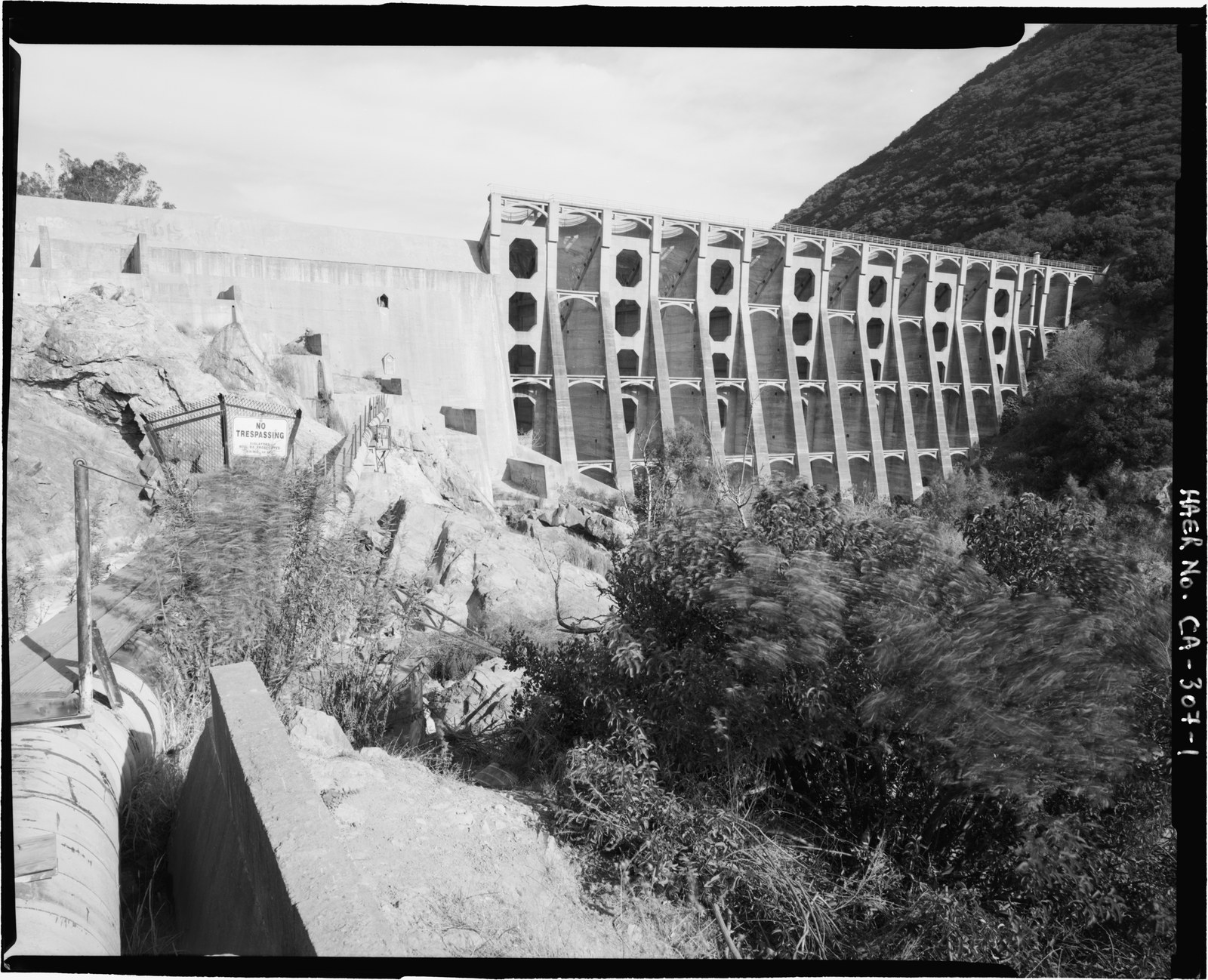 Lake Hodges Dam, a tall multiple arch concrete dam set between rock hillsides, with the concrete intake channel of the flume running along the ground in the foreground