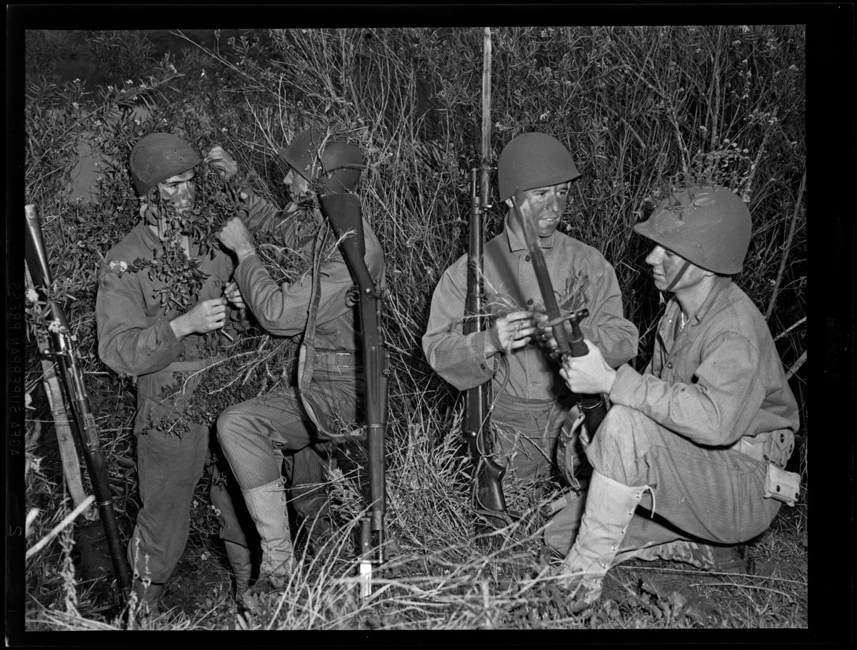 Four Marines in helmets crouch in thick brush tucking twigs into their gear, rifles with bayonets beside them
