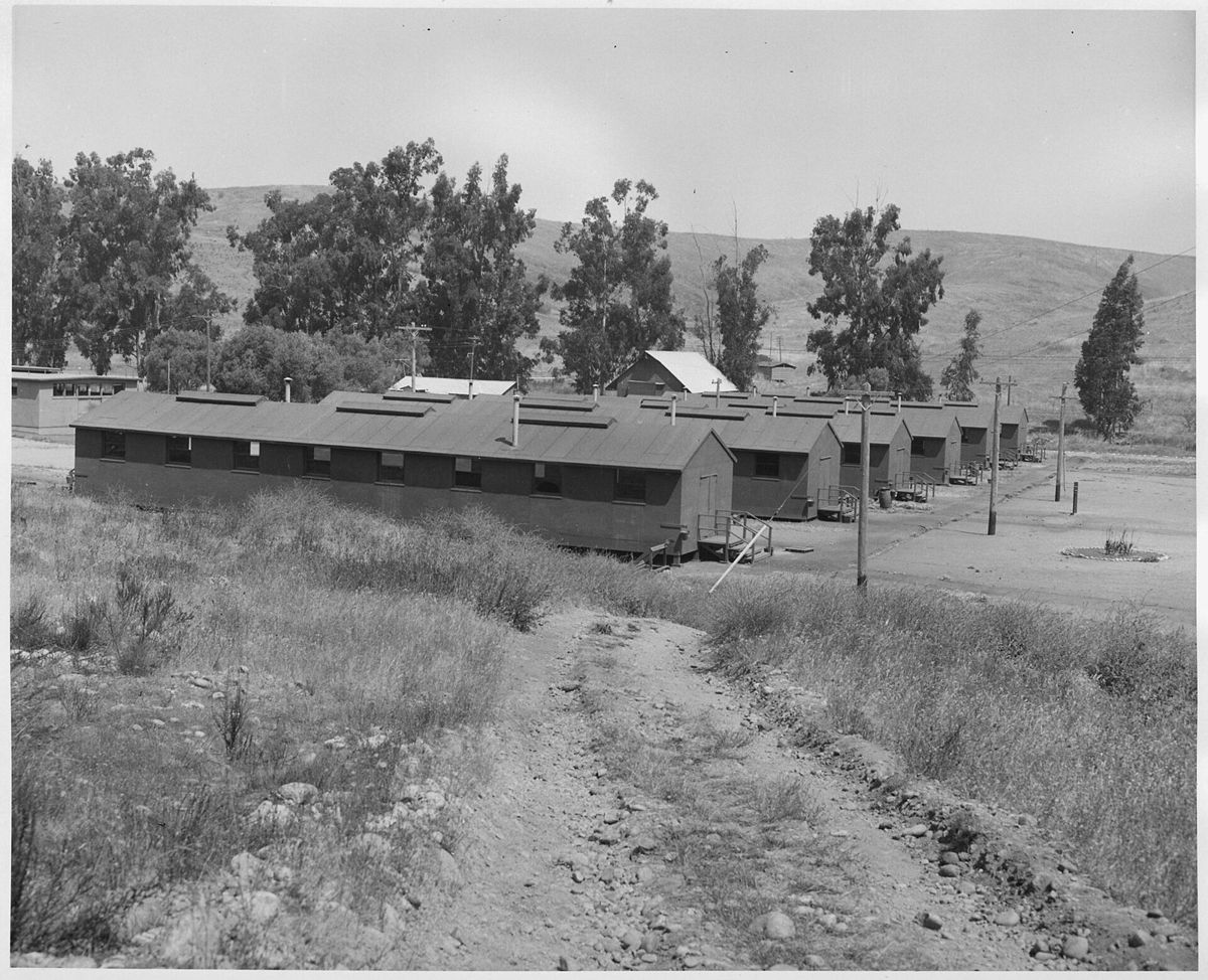 Rows of long single story wooden barracks on a dry mesa, a dirt track and wild grass in front and eucalyptus trees on the hill behind