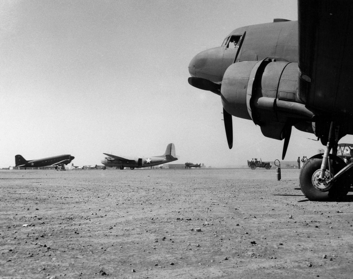 Twin engine transport planes parked on a wide flat dirt airfield, the nose and engine of one plane close in the foreground