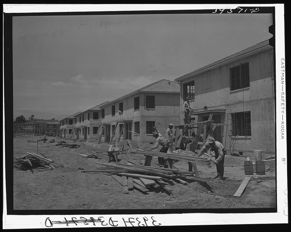 Workmen with lumber and sawhorses in the dirt in front of a row of new two story wood frame apartment buildings
