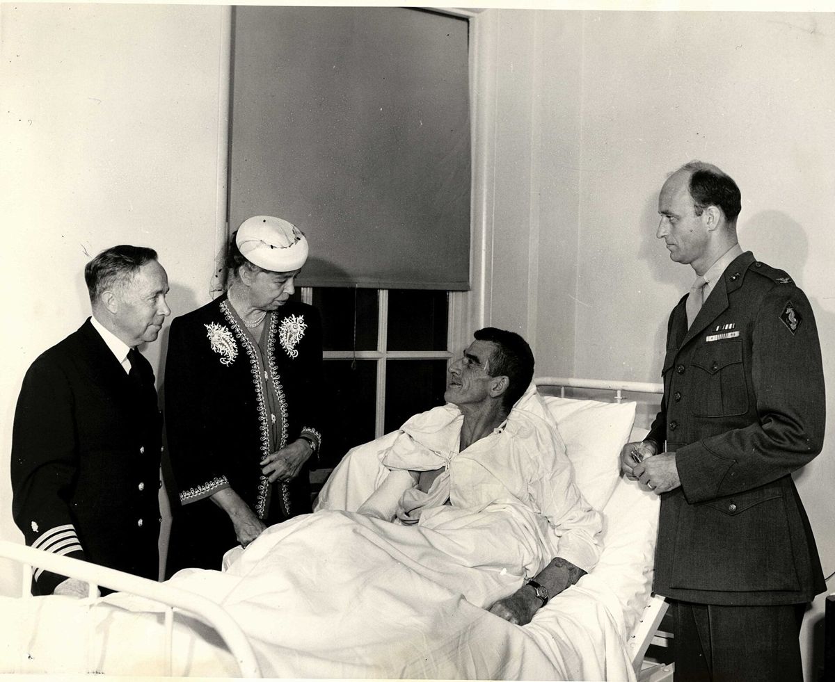 Eleanor Roosevelt in a hat stands beside a hospital bed where a wounded officer sits up, a Navy officer at left and a Marine colonel at right