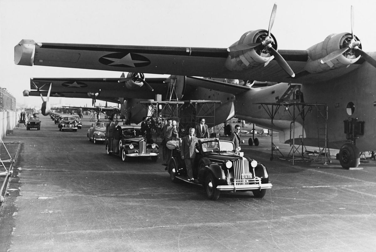 An open car carrying the President rolls down a long line of large four engine flying boats, workers and officials standing beside it