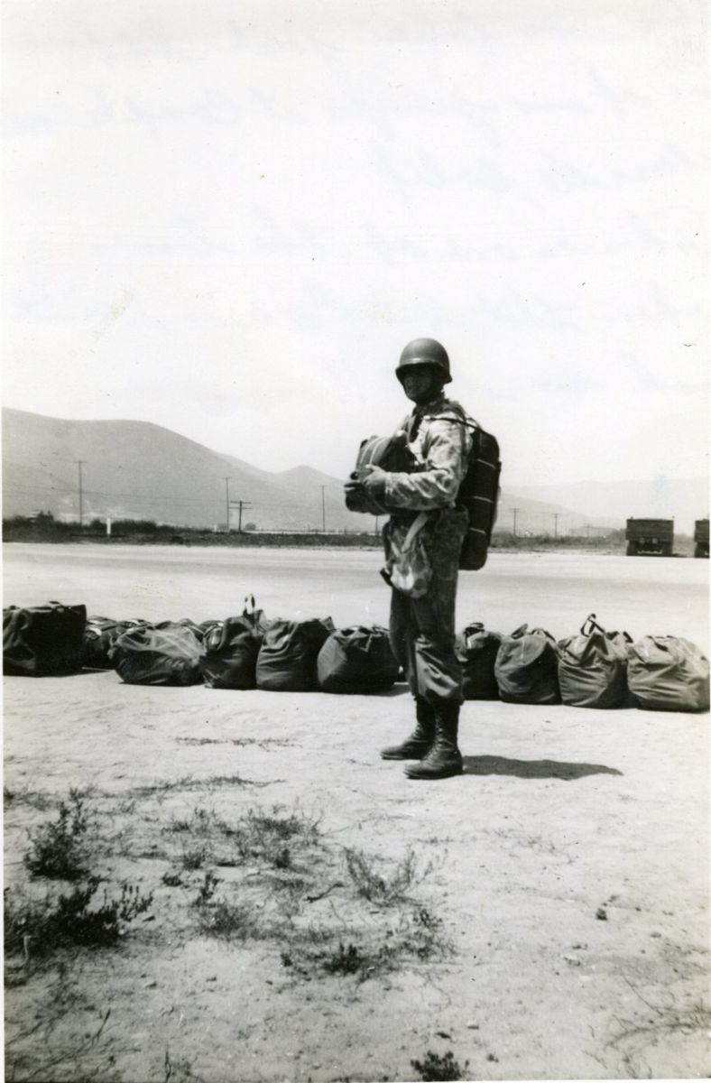 A Marine in a jump helmet and parachute harness standing on sand beside a row of packed parachute bags, mountains behind