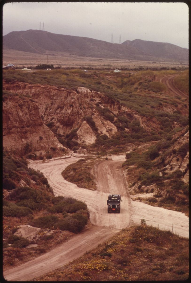 A military truck on a dirt road winding down through eroded coastal bluffs and wildflowers toward the sea, bare hills behind
