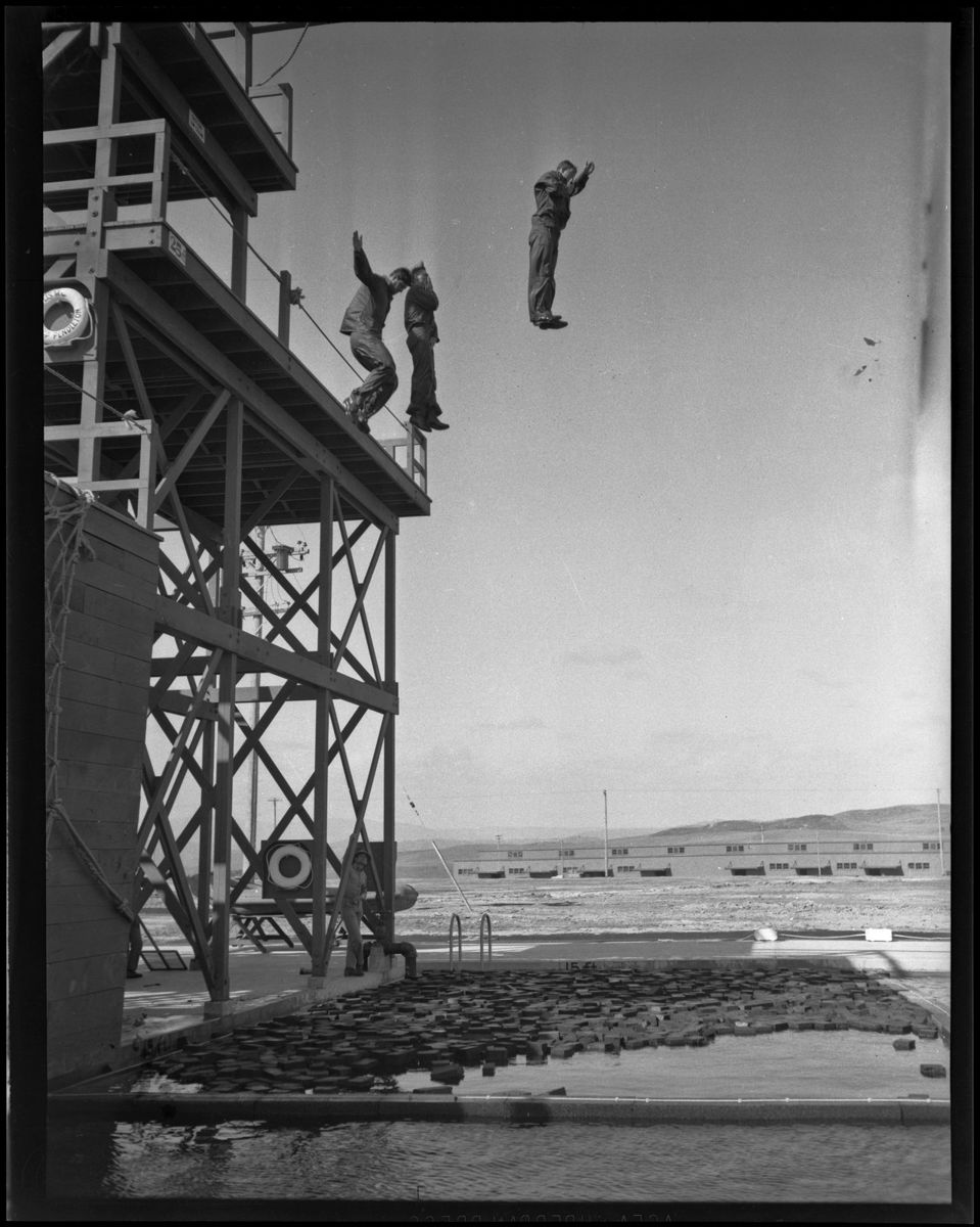 Two Marines in mid air dropping feet first from a tall wooden tower toward a pool covered in floating debris, hills behind