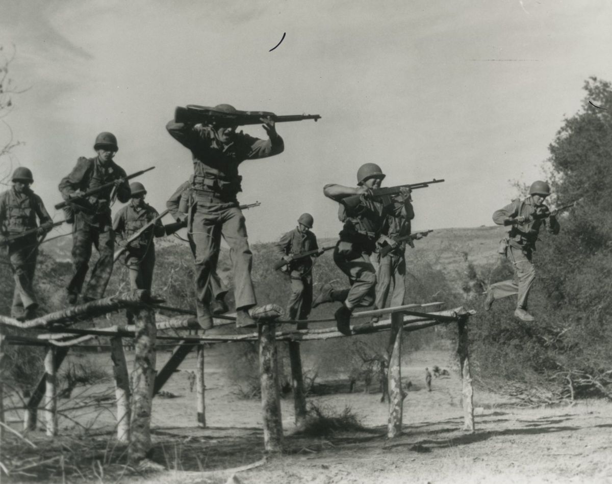 Marines in helmets carrying rifles run and leap along a raised course of narrow log beams over bare ground, brush and a hillside behind them
