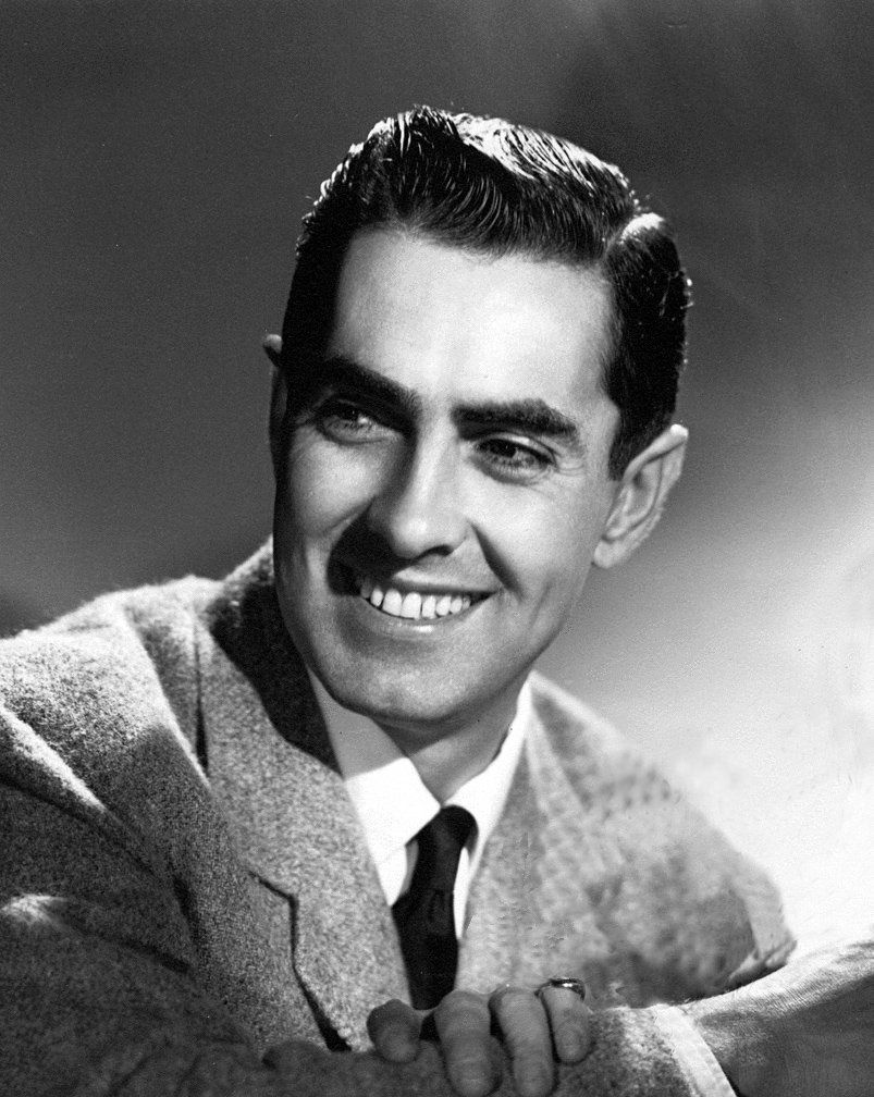 A studio portrait of a smiling dark haired man in a tweed jacket and tie