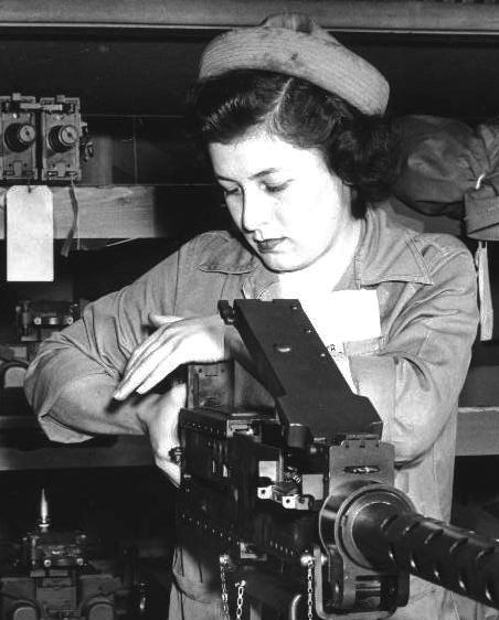 A young woman in a work uniform and cap bent over a heavy machine gun on a workbench