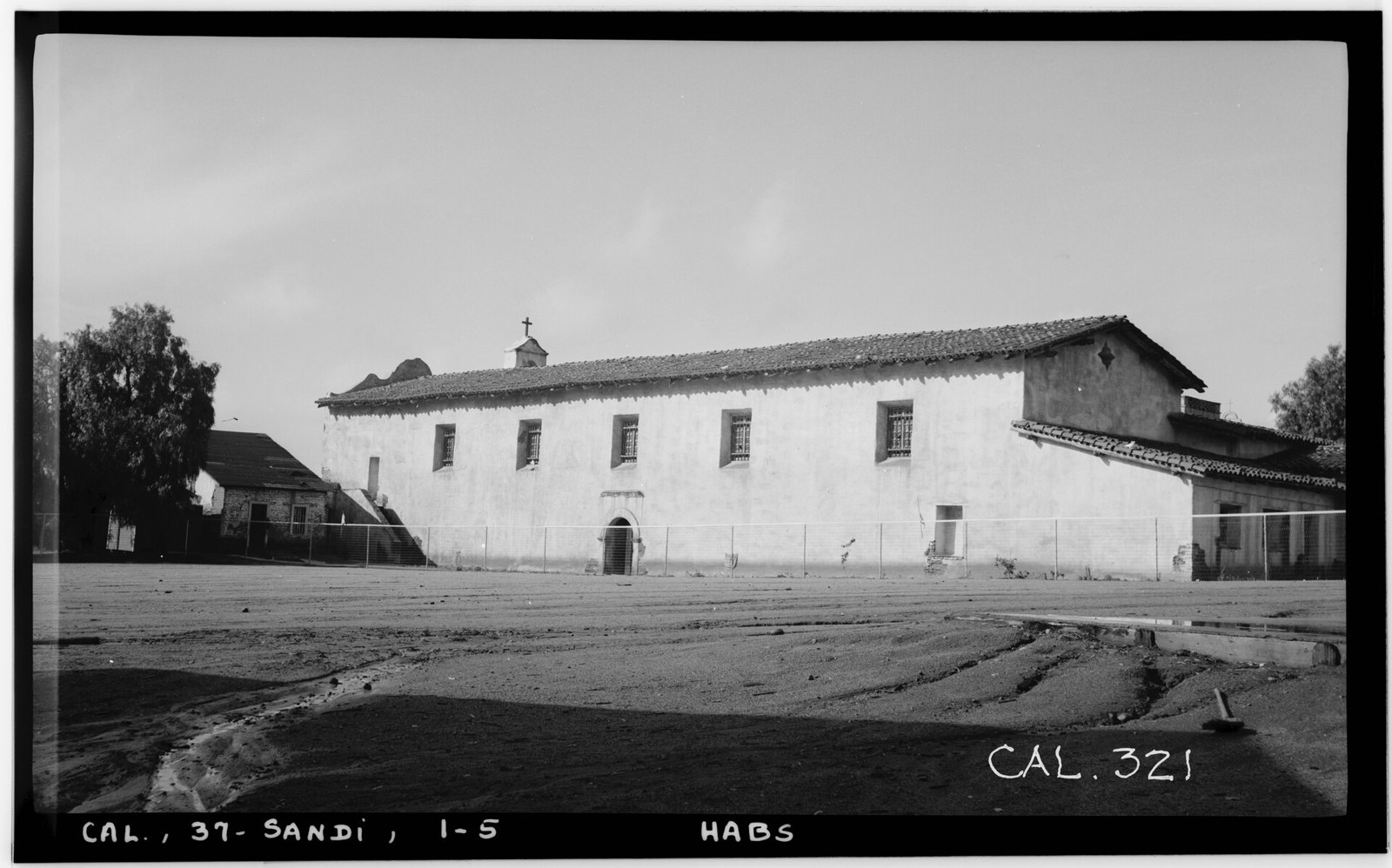 The long whitewashed front of the mission church with a bell wall at one end and a dirt yard in front