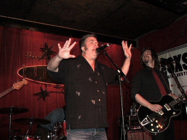 A man with both hands raised and mouth wide open, singing onstage in front of a deep red curtain, with band members behind him
