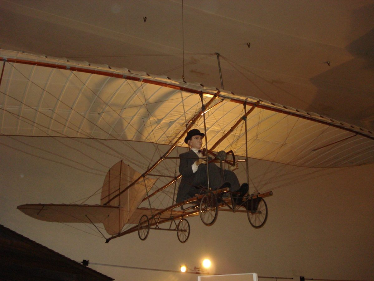 A glider with a long fabric wing and a seated mannequin pilot hanging from a museum ceiling