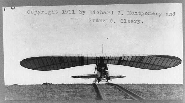 A monoplane glider with long curved wings sitting on a narrow rail track in an open field, a man seated under the wing