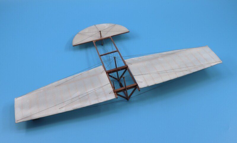 A small model glider with long curved fabric wings and a separate rounded tail on a wooden frame, against a blue background