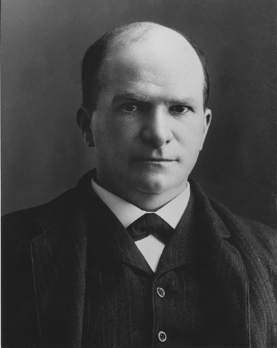 A studio portrait of a balding man with a mustache in a dark suit, high collar and bow tie