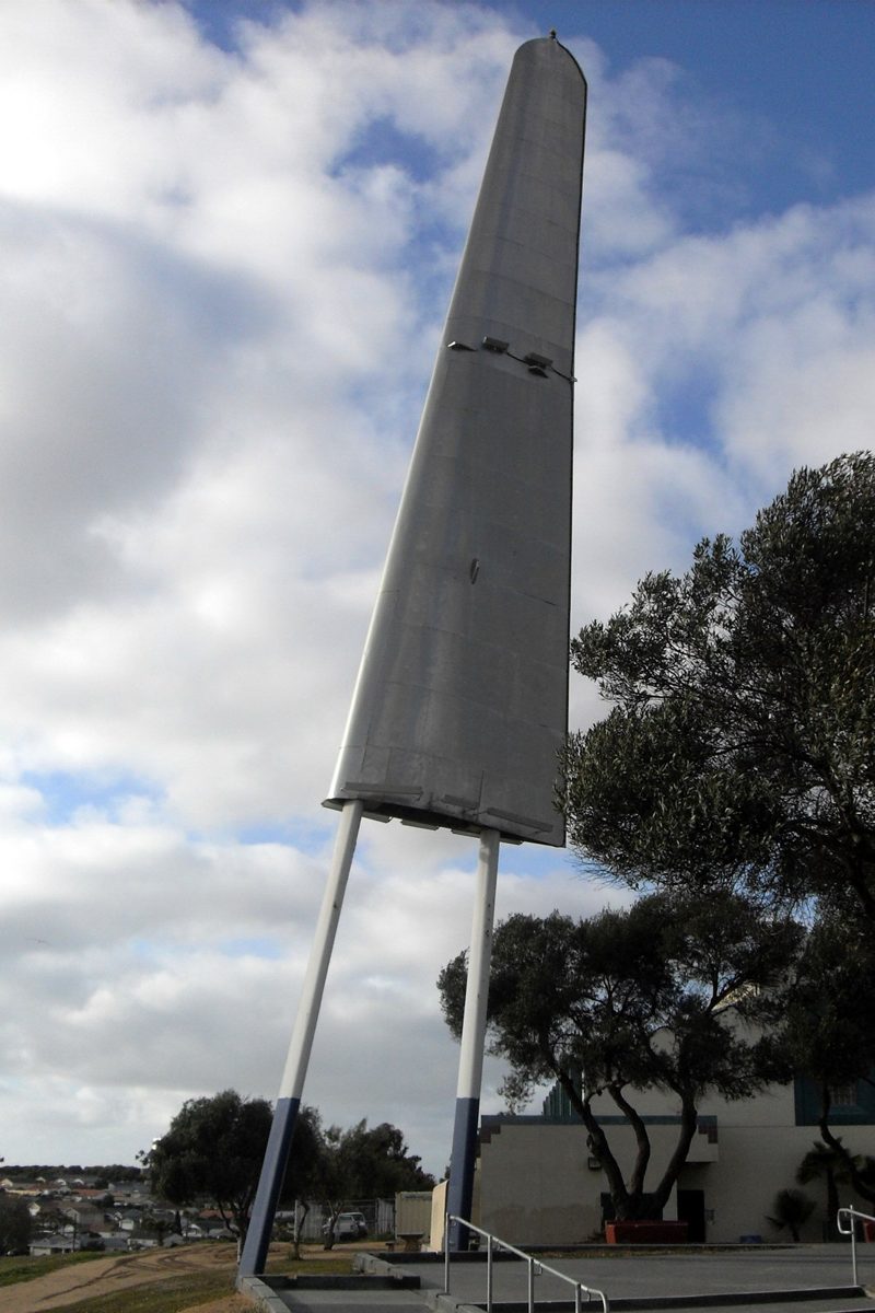 A tall silver aircraft wing mounted upright on steel legs in a park, trees and a low building behind it under a cloudy sky