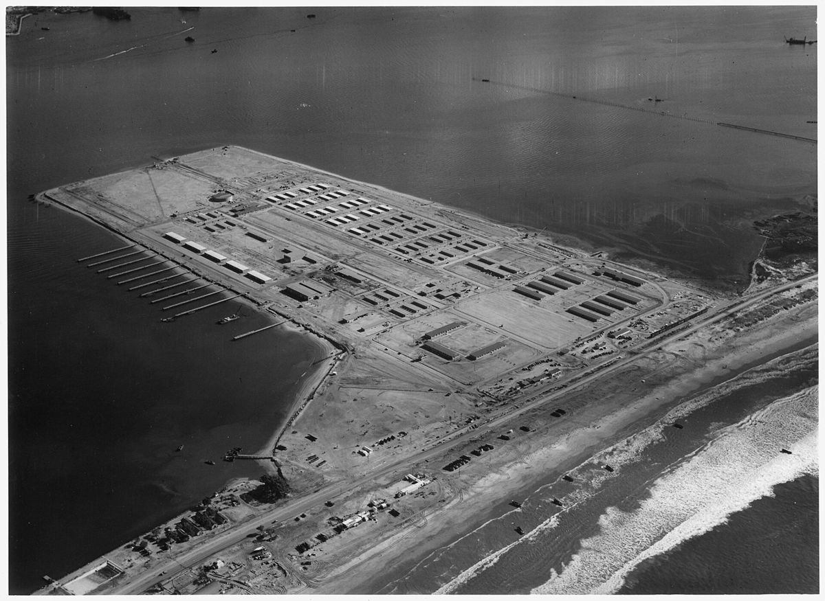 An aerial view of a new base of long buildings on a flat sand spit between the bay and the ocean, a highway running along it