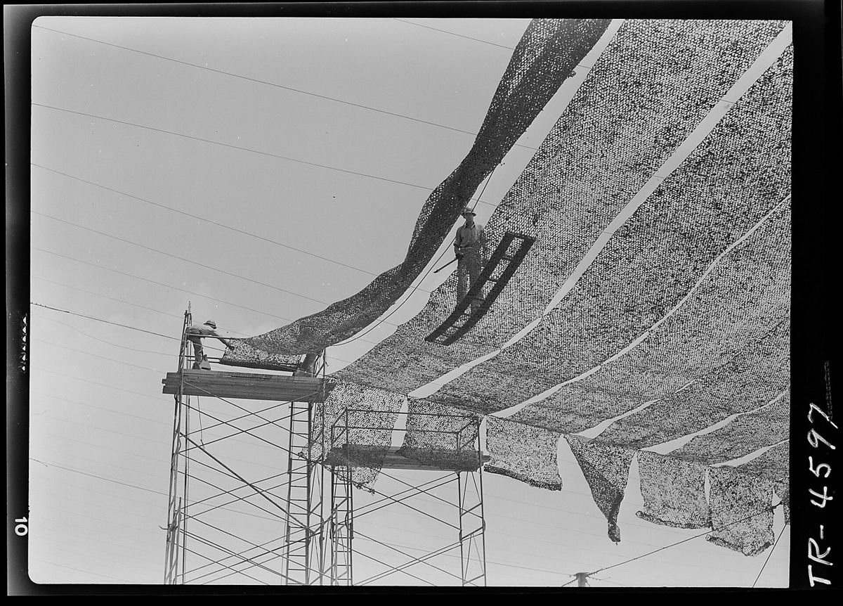 Two workmen high on steel scaffolding stretch patterned camouflage netting overhead