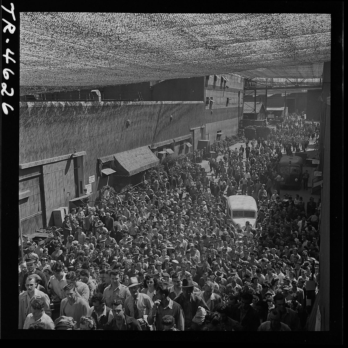 A street packed with thousands of workers walking toward the camera beneath a ceiling of patterned camouflage netting, a bus parked in the crowd