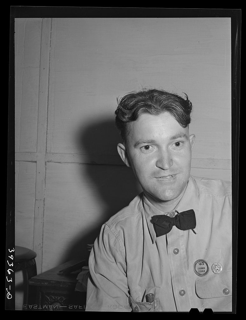 A young man in a work shirt and bow tie with a company badge on his chest looking at the camera