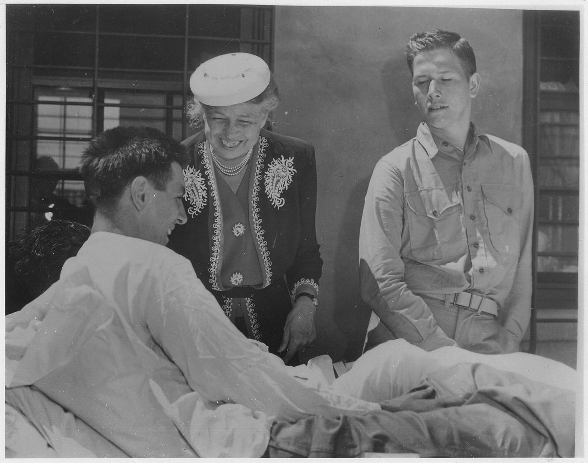 Eleanor Roosevelt in a white hat leans over to talk to a patient sitting in bed, a young sailor in hospital clothes standing beside them