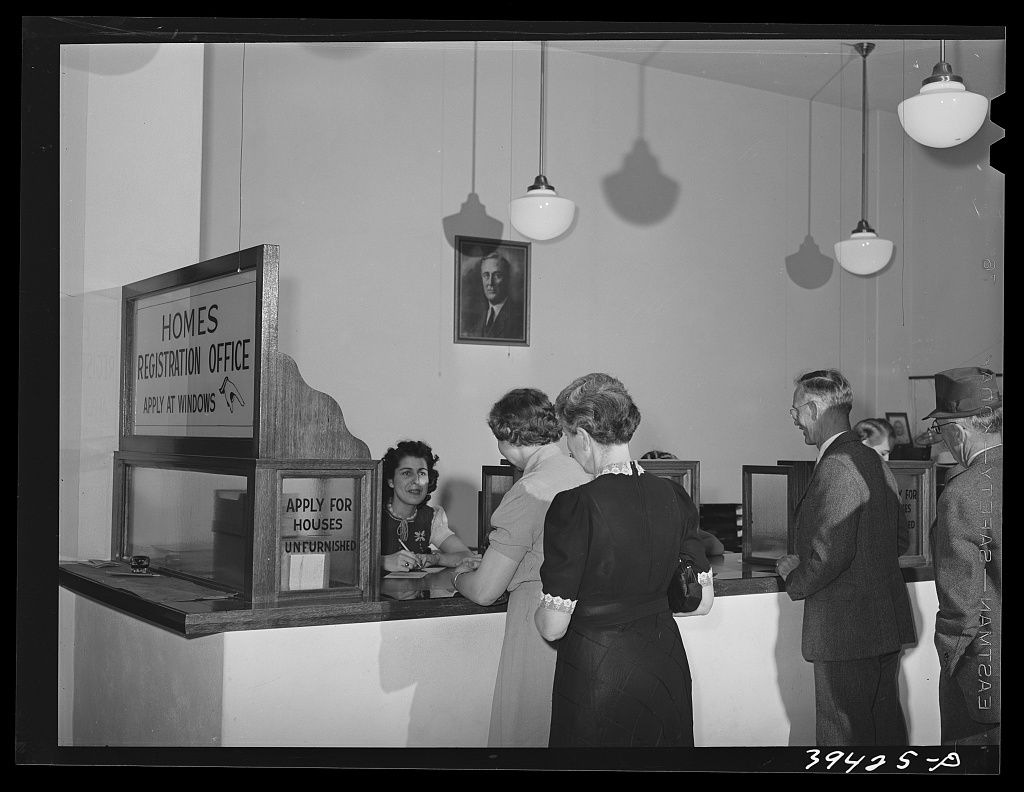 People at a counter under a sign reading Homes Registration Office, a clerk taking down a listing