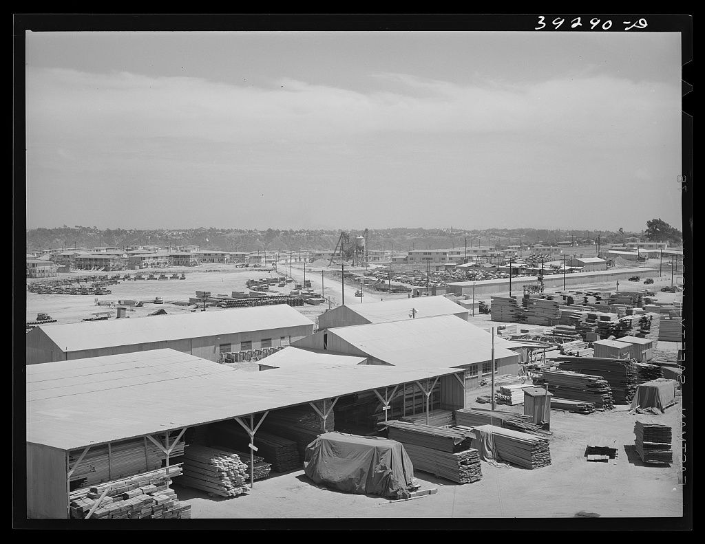 A wide view over sheds and stacks of building materials across a flat mesa, rows of houses in the distance