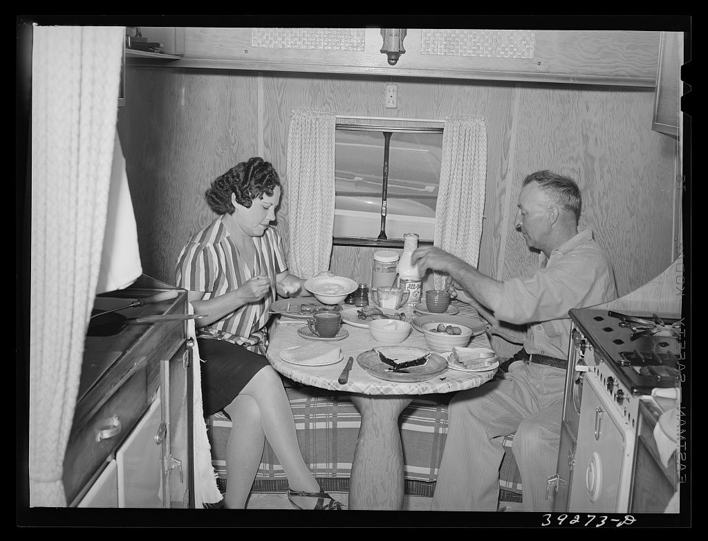 A man and a woman eating at a small round table inside a cramped trailer, a curtained window between them