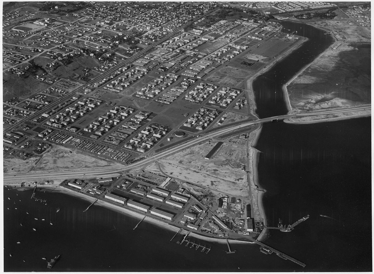 An aerial view of a large base of barracks and drill fields along a channel, neighborhoods spreading inland