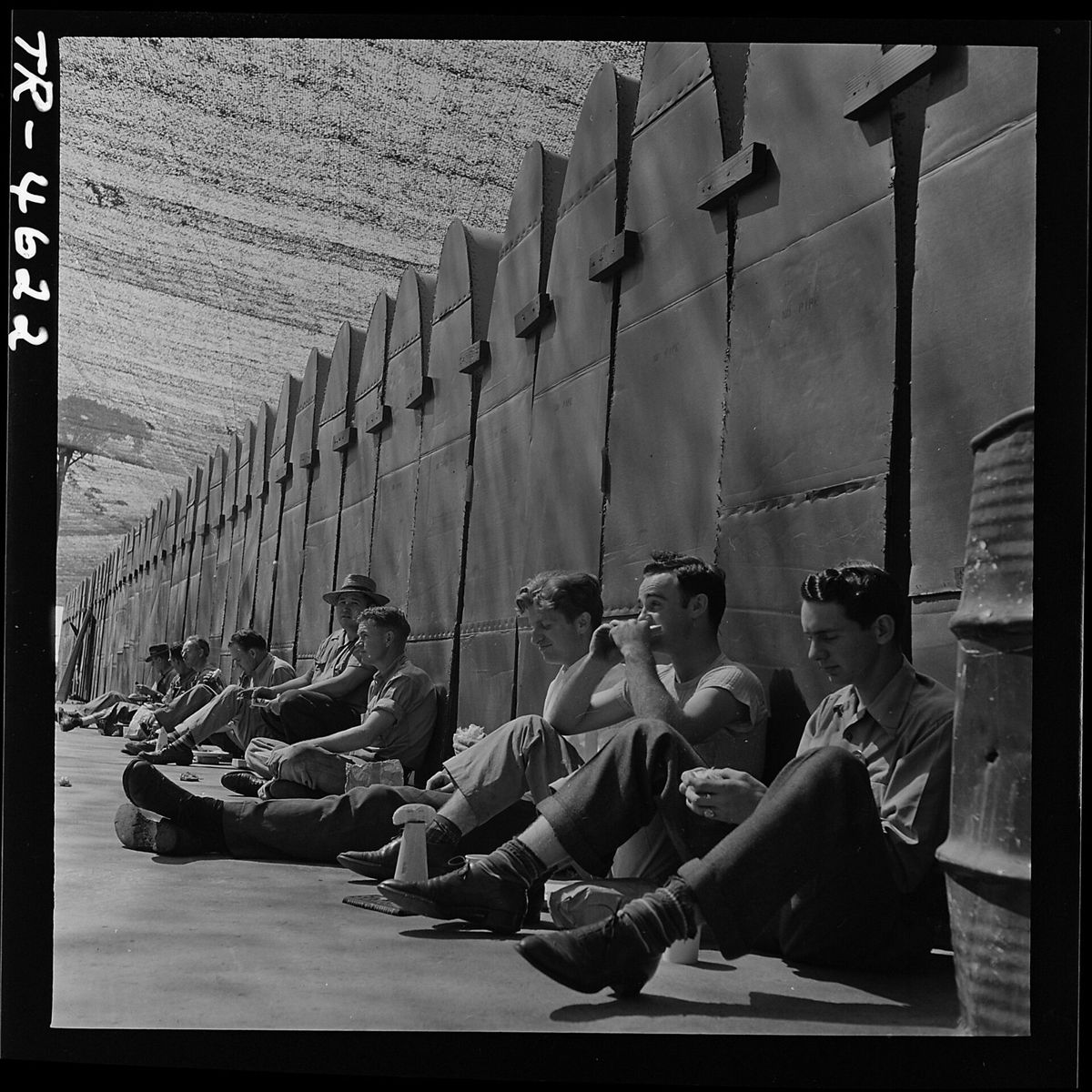 A line of workers sitting on the pavement eating lunch, their backs against a long row of tall upright aircraft wing panels