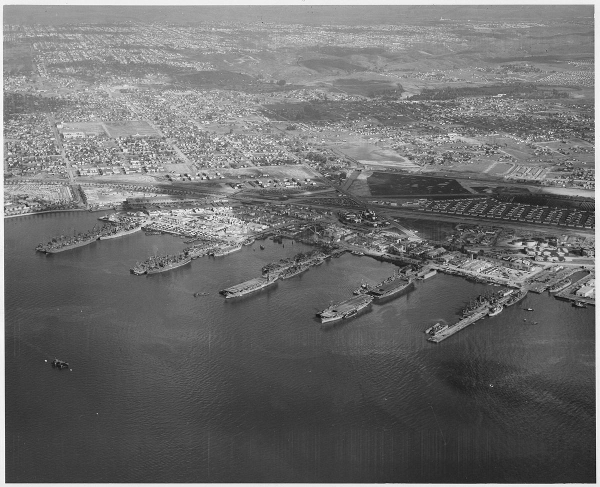 An aerial view of a row of large ships moored along piers on the bay, the city spread out behind