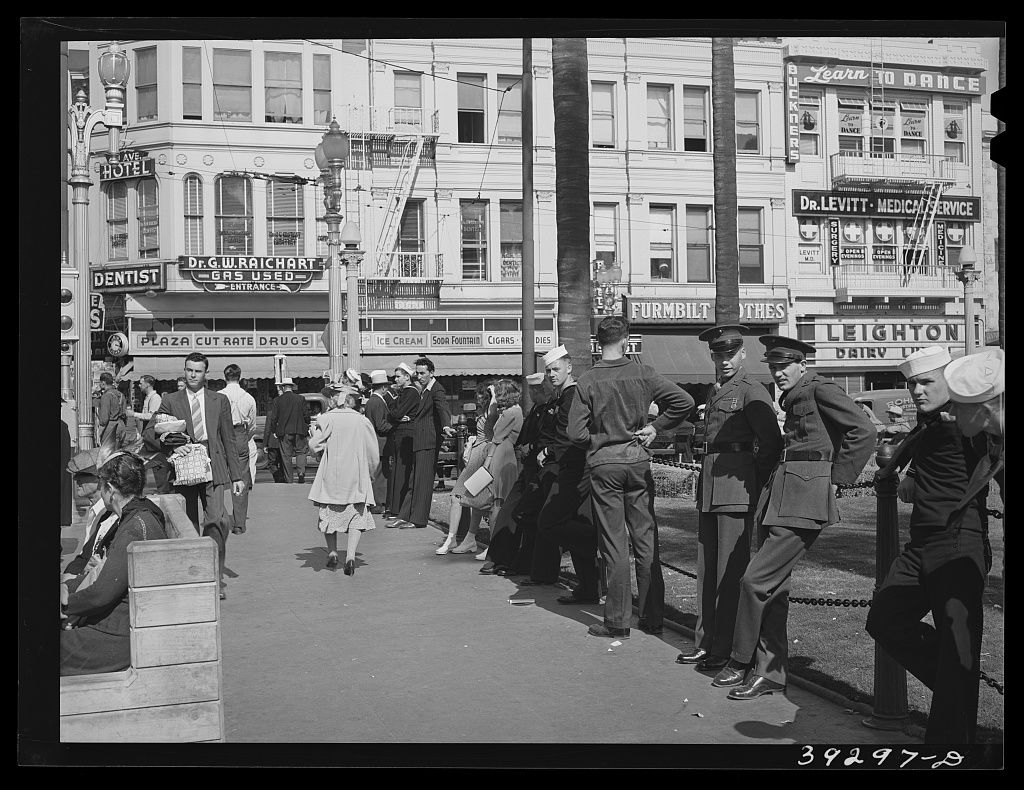 Sailors in white hats and Marines in caps lean on a low chain rail beside a plaza, storefronts, a hotel and a Leighton Dairy sign behind
