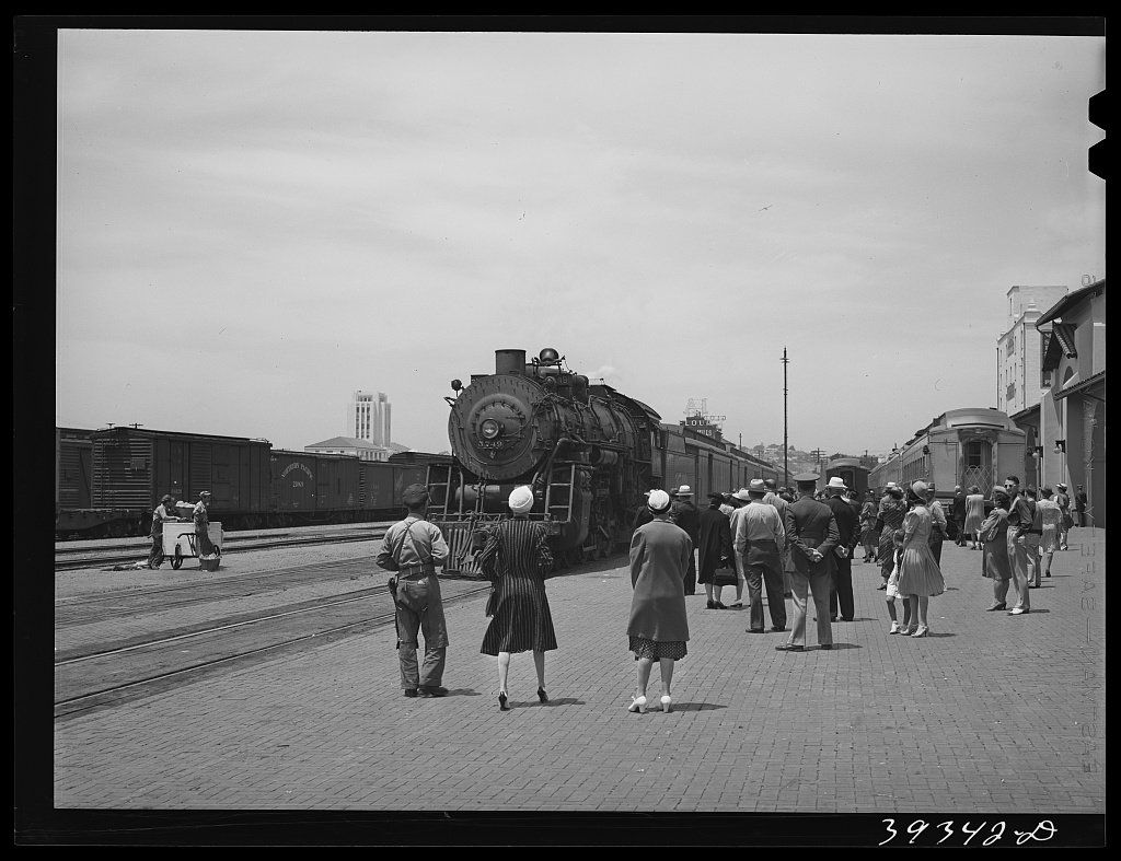 A steam locomotive at a station platform with travelers carrying bags walking along the brick paving