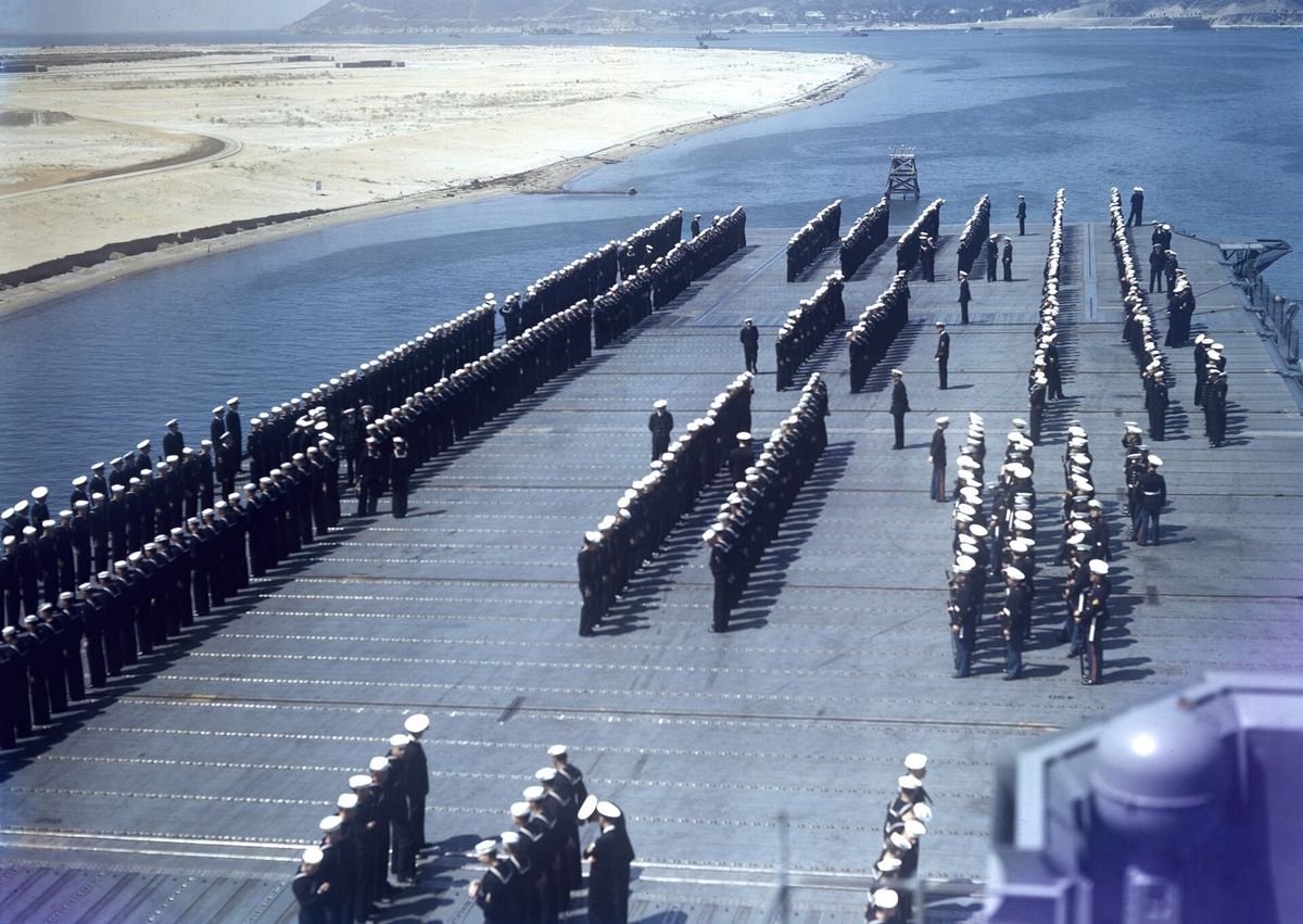 Rows of sailors in white uniforms lined up on a carrier flight deck, the bay and a sandy shore beyond