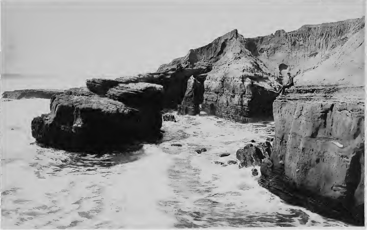 The sandstone shelf and sea cliffs at Ocean Beach photographed in 1887, surf running over flat rock in the foreground and a man in a hat seated on the cliff edge at upper right