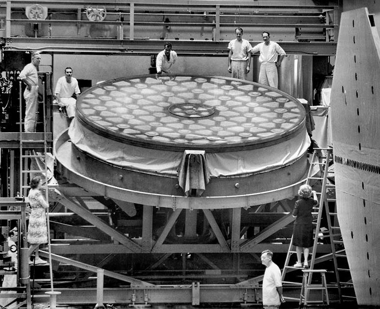 Seven people standing around the 200-inch telescope mirror lying face up on its grinding machine in the Caltech optical shop in December 1945, the honeycomb pattern visible through the glass