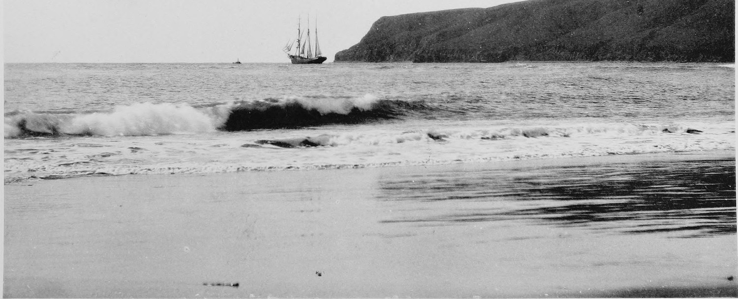 A sailing ship lying off a high dark headland, a wave breaking across the foreground and wet sand beyond