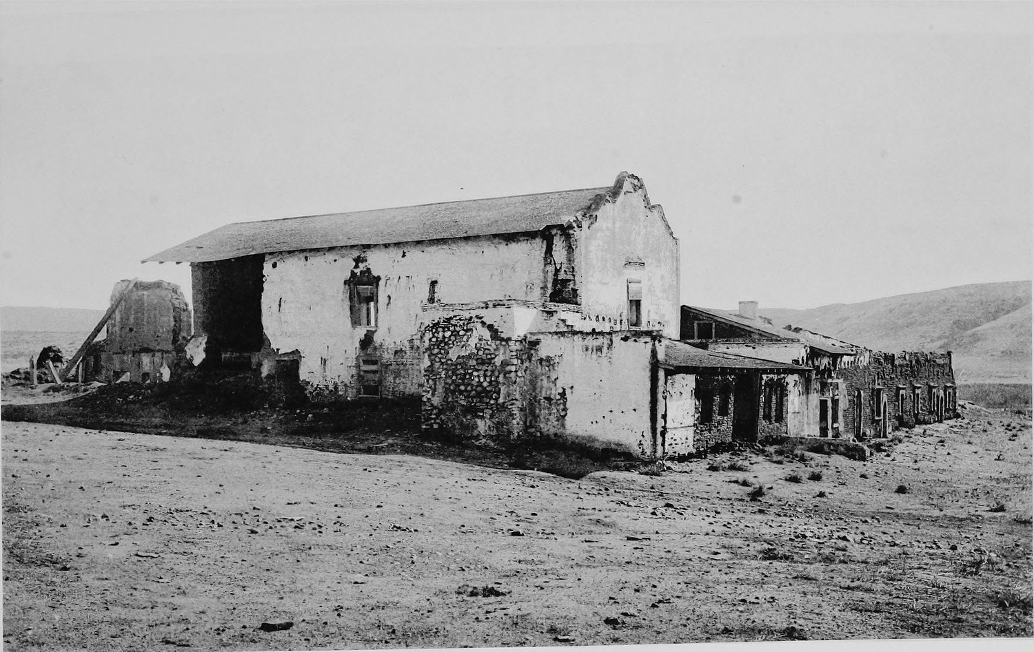 A long adobe mission church with a gabled roof and a crumbling ruined facade, standing alone on open dry ground