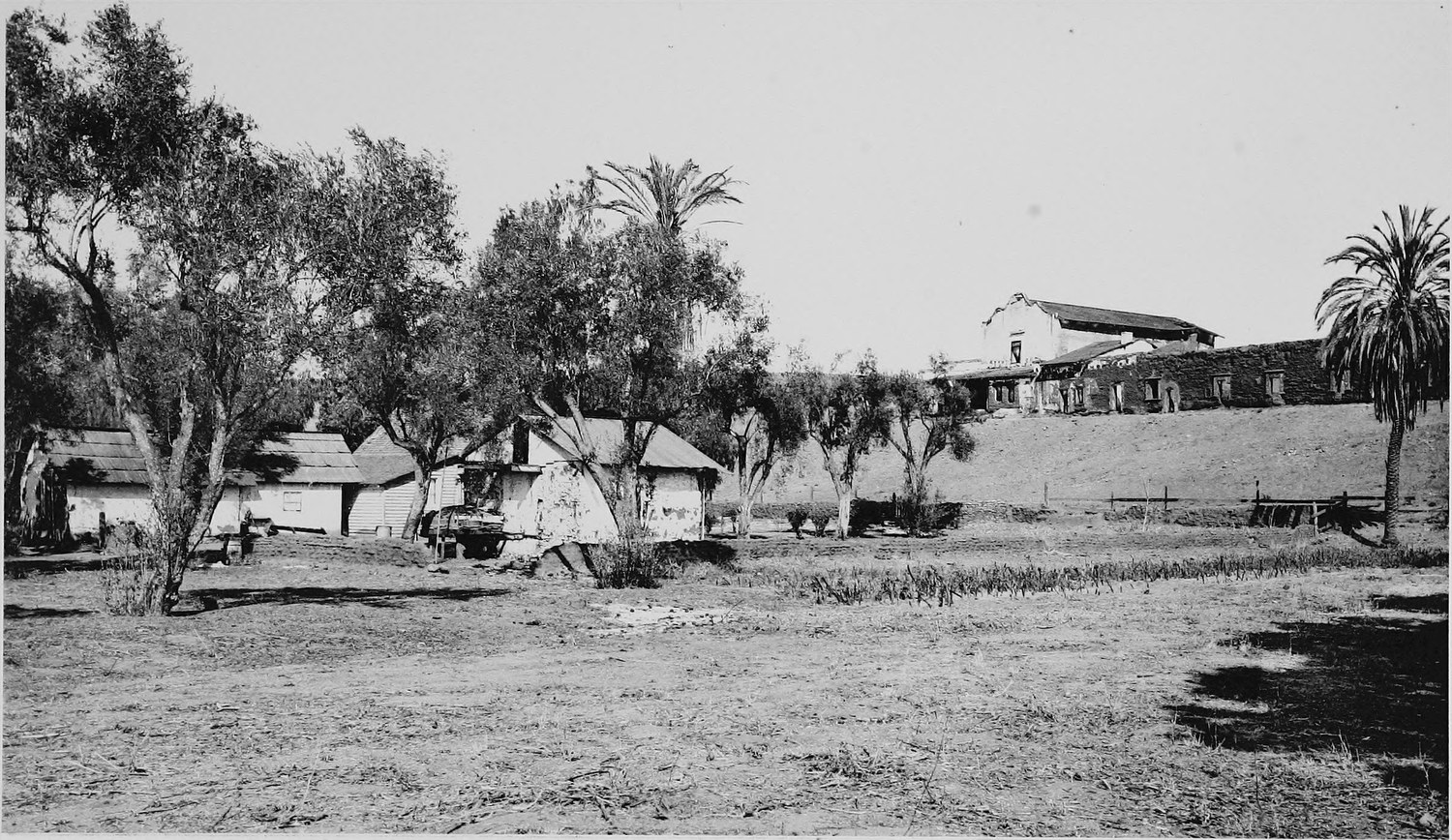 A low adobe mission and its outbuildings seen across a dry field, old trees and palms around the buildings
