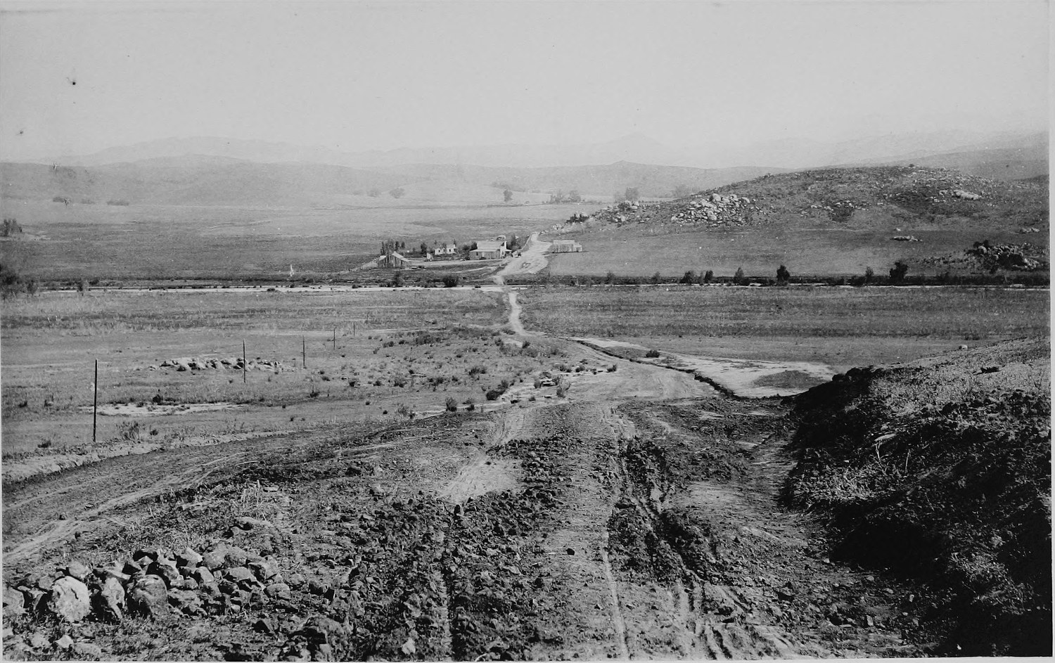 A rutted dirt road running down a bare treeless valley toward distant hills, a few small buildings in the middle distance